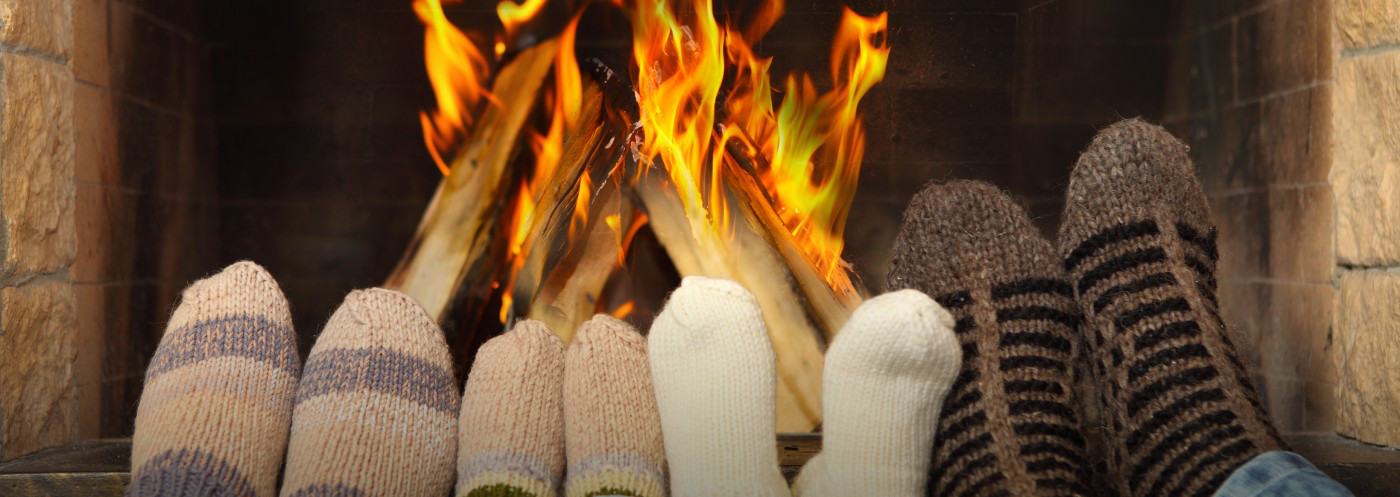 Four people keep warm by a fireplace. They are wearing socks and the their feet by the fire to enjoy the warmth.