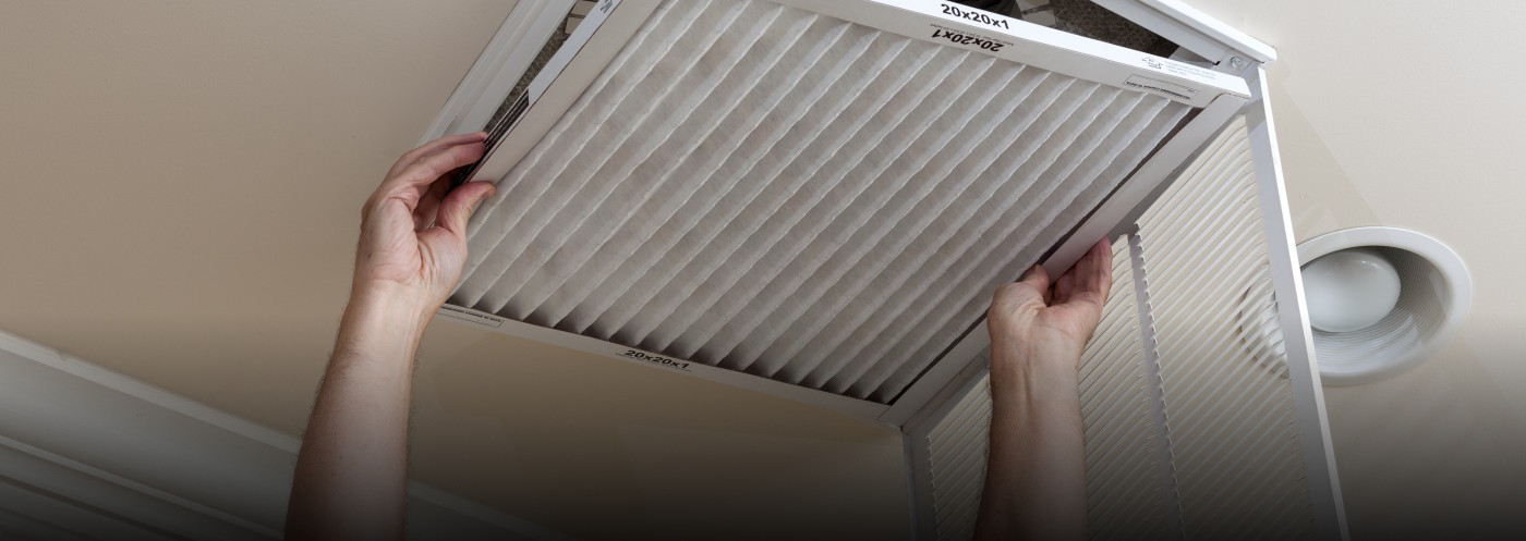 A person reaches up to replace an HVAC system filter. This is part of basic HVAC maintenance and will help the system run more efficiently.